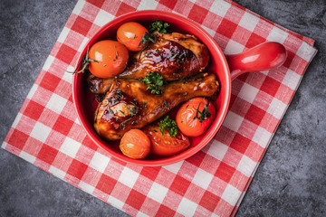 baked chicken legs in a barbecue sauce with tomatoes in a red frying pan, top view