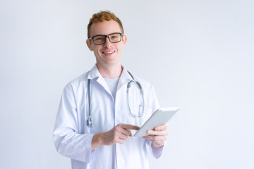 Happy young male doctor using tablet computer. Guy wearing white coat and standing. Medicine and technology concept. Isolated front view on white background.