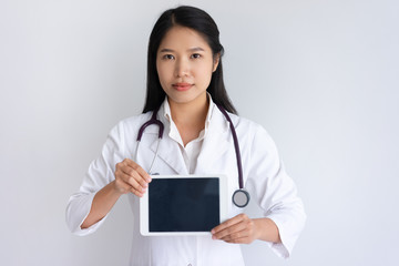 Serious female doctor showing tablet computer screen. Pretty young Asian woman wearing white coat and standing. Healthcare and technology concept. Isolated front view on white background.