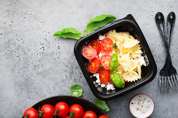 Meal prep. Black lunch boxes. Pasta bows, cherry tomatoes, young cheese, salt, pepper, basil. On a dark background Top view.