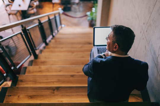 Middle-aged businessman dressed smart casual sitting on the stairs indoors and using laptop for work.