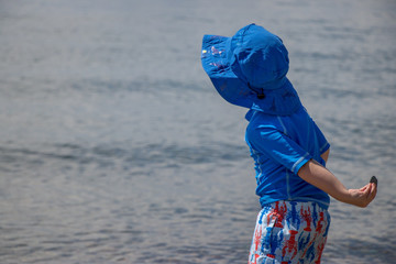 toddler boy wearing sun protective clothing and hat throws rock in ocean