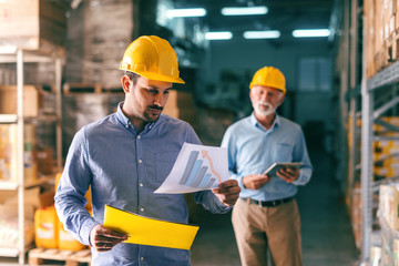 Young Caucasian man with serious face wearing helmet and looking at chart while standing in warehouse.