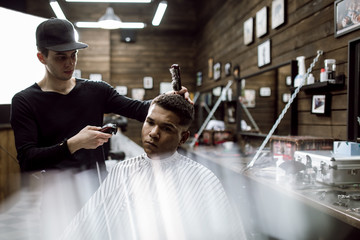 The fashion barber in black clothes makes a razor cut hair for a black-haired man sitting in the...