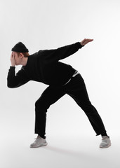 Young freestyle dancer dressed in black jeans, sweatshirt, hat and gray sneakers is dancing  closing his face with his hand in the studio on the white background
