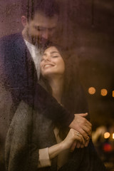 Romantic loving guy hugs his happy girlfriend standing behind a wet window with lights.