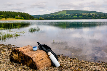 Hiker backpack and insulated flask set on a tree stump at the edge of the water. Keeping warm when hiking on a cold winter day. Lake side landscape at Blessington Lake in Wicklow Mountains, Ireland. 