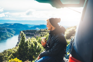 hipster tourist hold in hands mug of hot drink, lonely guy enjoy sun flare mountain in auto, happy traveler drink cup of tea on nature, vacation weekend concept on panoramic landscape