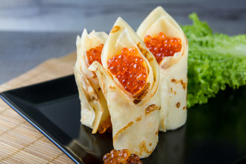 horizontal photo of the black plate with pancakes filled by red caviar and salad leaves 