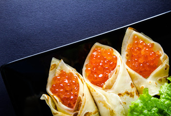 horizontal photo of the black plate with pancakes filled by red caviar and salad leaves 