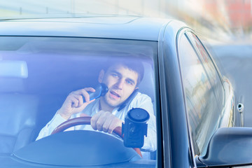 A young guy is a black car driver talking on a mobile phone / smartphone, drinking tea, coffee and shaving with an electric razor. The concept of not safe driving