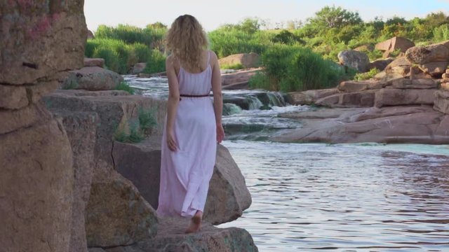 slim tall model with blond curly flowing hair slowly walks barefoot on stones, a forest nymph or river fairy in a long pink summer light dress with a flying hem, no face, bright juicy colors
