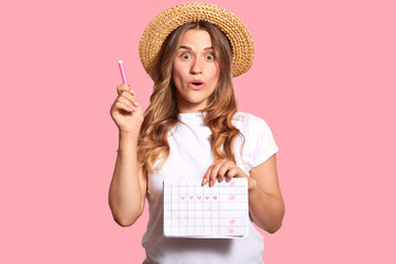 Waist up shot of amazed beautiful woman holds pen and menstrual calendar, shows five day marked with hearts, isolated over pink background. Attractive girl checks her periods, stands indoor.