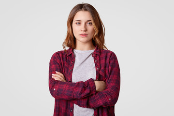 Indoor shot of serious self assured teenage girl keeps hands crossed, wears checkered shirt, looks...