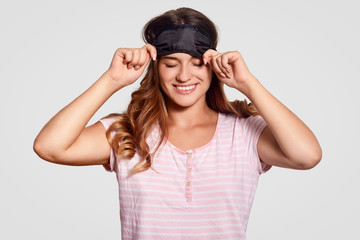 Fototapeta na wymiar Glad optimistic Caucasian woman wears eye mask, has broad smile, dressed in pyjamas, has happy expression, sees pleasant dreams, models against white background. People, wellness, rest concept
