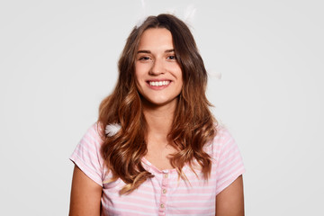 Fototapeta premium Bed time concept. Cheerful young woman with toothy smile, has feathers on head, wears pyjamas, ready for good rest, isolated over white background, expresses positive feelings, models indoor