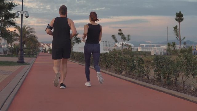 Pair of sporty guy and girl are jogging in park area in evening, back view