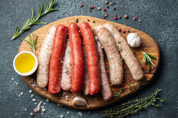 Assorted fresh sausages with thyme, rosemary, olive oil and garlic on a wooden chopping Board on a dark concrete table. Ready to cook sausages for dinner on black background. Top view with copy space