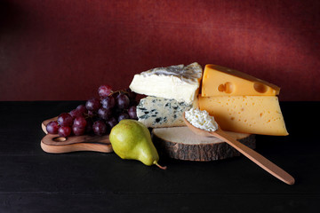 Cheese, grapes and pear on a wooden stand. Different types of cheese and fruit on a dark background. Camembert, blue cheese and cottage cheese. Dairy products and a bunch of grapes