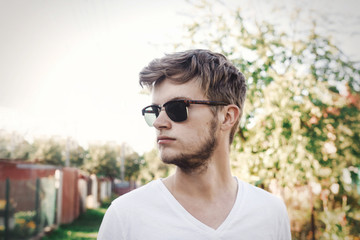 Stylish hipster man in sunglasses posing outdoors in sunny countryside. Handsome bearded man relaxing in evening summer city. Vacation and travel