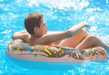 Cute child boy on funny inflatable donut float ring in swimming pool. Teenager learning to swim, has fun in outdoor pool at tropic resort. Water toys for kids. Healthy sport activity, vacation 