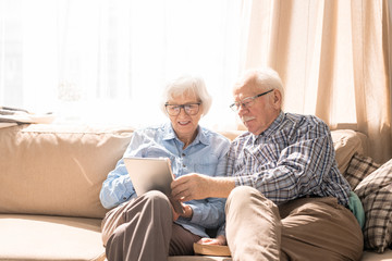 Portrait of modern senior couple using digital tablet sitting on couch at home in sunlight copy...