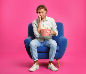 Fototapeta premium Emotional man with popcorn sitting in armchair during cinema show on color background