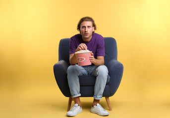 Emotional man with popcorn sitting in armchair during cinema show on color background