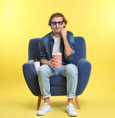 Fototapeta premium Emotional man with 3D glasses, popcorn and beverage sitting in armchair during cinema show on color background