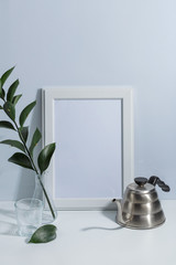 Mock up white frame, coffee pot and green twigs in vase on book shelf or desk. White colors. White-blue colors. Minimalistic concept.