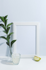 Mock up white frame, green twigs in vase, piece of apple and glass of limonade on book shelf or desk. White colors. White-blue colors. Minimalistic concept.