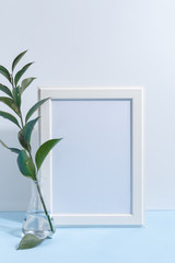 Mock up white frame and green twigs in vase on book shelf or desk. White colors. White-blue colors. Minimalistic concept.