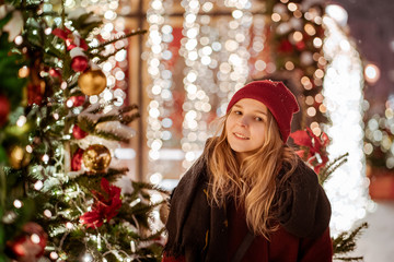 Outdoor close up portrait of young beautiful girl with long hair wearing red hat, posing in street with christmas ornament. Christmas, winter holidays concept.