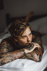 Young handsome tattooed man just woke up and laying in bed in the bedroom.