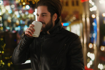 Young guy with beard is drinking warming drink from paper cup. Portrait on night street of city. Bokeh from festive lights.