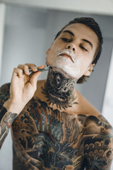 a young handsome tattooed man in the morning shaves in front of a mirror, a razor in his hands, a...