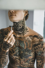 a young handsome tattooed man in the morning shaves in front of a mirror, a razor in his hands, a...