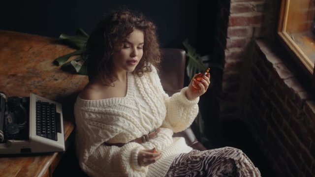 Young beautiful confident woman holding, sniffing luxury perfume in glass bottle. Model wearing warm winter knitted sweater.