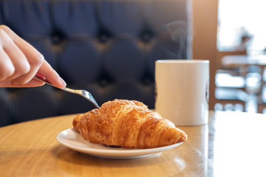 Closeup image of hands cutting a piece of croissant by fork for breakfast with coffee cup on wooden table