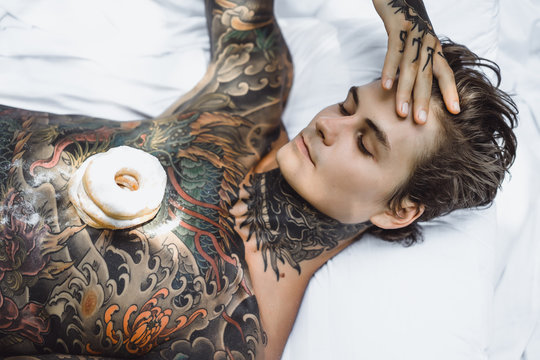 young handsome tattooed man having breakfast in bed in the open air outdoors in the garden, donuts and coffee for breakfast. - Powered by Adobe