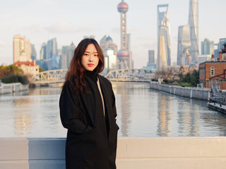 Fototapeta premium Beautiful young brunette woman in black clothes smiling at camera with blur Shanghai Bund landmark buildings background with dusk light. Emotions, people, beauty, travel and lifestyle concept.