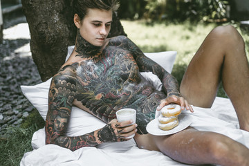 young handsome tattooed man having breakfast in bed in the open air outdoors in the garden, donuts...