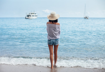 Back view of young woman in hat enjoy sea and boats from the beach