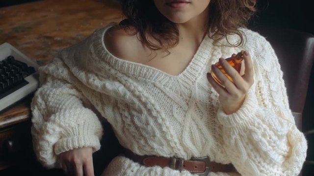 Young beautiful fashionable lady holding, using luxury perfume in glass bottle. Model wearing warm winter knitted sweater.