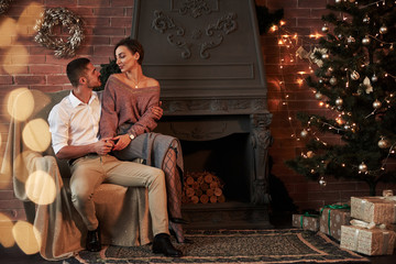 Beautiful young couple on the chair hugging in luxury room that is decorated holiday tree and garlands