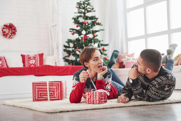 Obraz na płótnie Canvas Girl is excited what kind of gift inside the box. Lovely young couple lying on the living room with green holiday tree at background