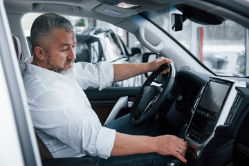 Buying and testing new automobile. Senior businessman in official clothes sits in a luxury car and pushing the buttons on the music player