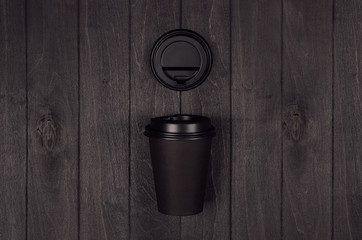Coffee packing mockup - black paper cup and blank cap on dark black wood board with copy space, coffee shop interior. Modern elegant concept for branding identity, advertising, design.