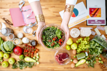 Dietitian holding cooked salad above the table full of various healthy products and drawings on the topic of healthy eating