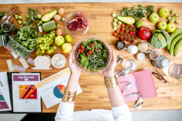 Dietitian holding cooked salad above the table full of various healthy products and drawings on the...